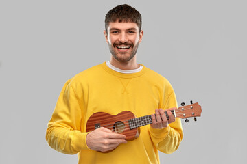 music and people concept - smiling young man in yellow sweatshirt playing ukulele guitar over grey background
