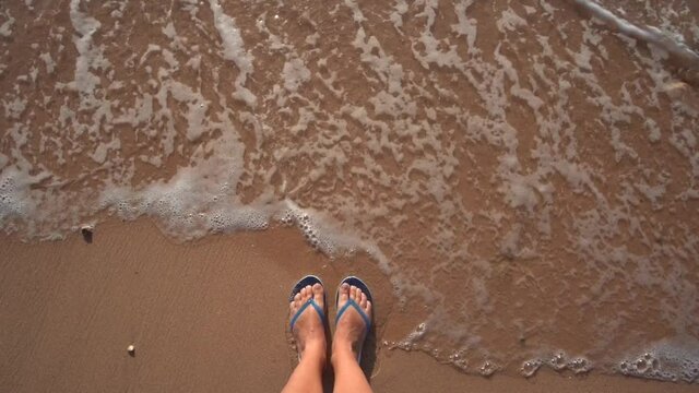 Closeup top view video of legs of woman enjoying summer vacations standing on sandy sea beach. Close-up view of female naked barefoot legs wearing flip-flops isolated on sand background.