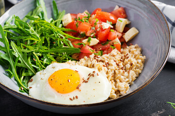 Breakfast oatmeal porridge with fried egg, tomato salad, arugula and flax seeds. Healthy balanced food.