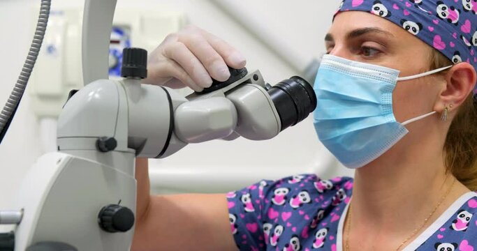 Woman dentist (dental surgeon), wearing medical mask and gloves, adjust her microscope eyepiece at dental clinic. Medicine, dentistry and health care concept with specialized dental equipment.
