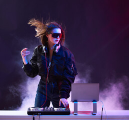Stylish girl DJ in glasses headphones and a bomber mixes music dancing at a party in the neon light...