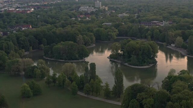 Aerial drone shot, zoom out over Munich. The early sunrise view at a lake in Munichs famous Englischer Garten out to the horizon line with the overview to the north of the bavarian capital.