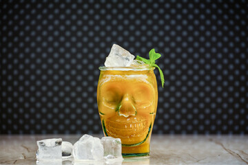 Peach juice in a skull-shaped glass at a marble board with pieces of ice. Fresh peach juice, ice and an unusual mug in shape of a head. Halloween zombie cocktail in a glass skull-shaped.