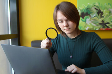 A young beautiful girl of European appearance in a blue jacket sitting in a cafe and working on a laptop. Looks at a computer display through a magnifier.