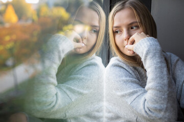 Depressed/anxious young woman sitting by a large window, feeling blue, sad, uncertain - obrazy, fototapety, plakaty