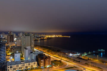 Fototapeta na wymiar Long exposure panoramic night view of the coastline at a holiday resort in Punta Del Este, Uruguay: the quiet empty beach, the calm water, the car lights passing and the illuminated harbor at the back