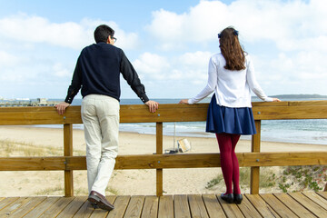 A well dressed young couple from behind on a wooden deck pier looking down at the empty beach and...