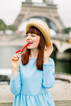 Young charming red haired laughing woman, wearing straw hat and elegant blue dress, posing for photo, biting red lollipop, while standing on Eiffel tower background and river Seine