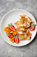 Crepes with cream, fresh strawberries, peach, top view on white plate. Russian thin blinis with cottage cheese on light concrete background.