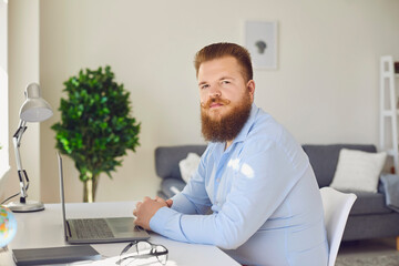 Online work at home. A serious fat bearded man is looking at the camera in the workplace at home.