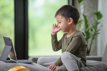 Asian boy looking at the tablet screen attentively. overstimulated children concept. Too much screen time. 3 years boy watching videos while tv is working.Smartphone, Internet addiction concept.