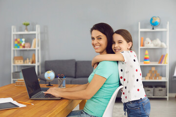 Lovely girl hugging her mom while she working online via laptop at home. Cute child embracing her...