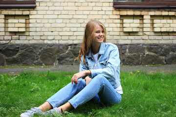 Fototapeta premium Cute positive fair-haired girl dressed in jeans with a smile on her face sits on the grass near the university.