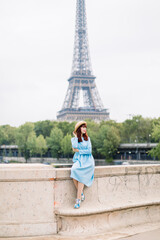 Young woman in blue dress enjoying beautiful landscape view on the Paris and Seine river, while sitting on the bridge on the background of Eiffel tower
