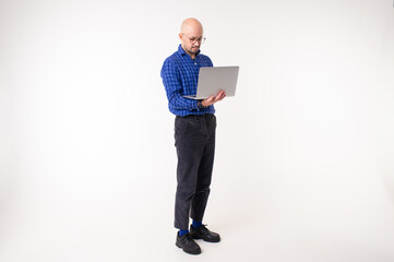 Handsome caucasian man without hair in blue shirt, black trousers and blue socks works with laptop isolated on white background