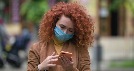 Beautiful young red-haired caucasian woman in medical mask using smartphone social media