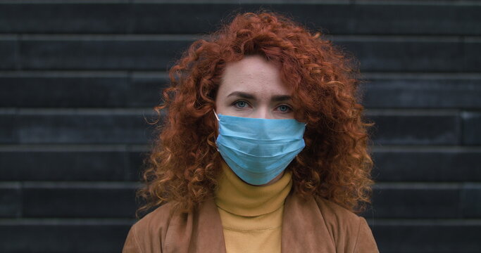 Beautiful young red-haired caucasian woman puts on a medical mask looking into the camera