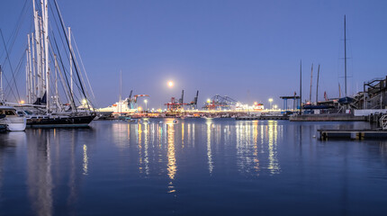 Fototapeta na wymiar harbor docked sailboats and cargo port cranes full Moon sky and water reflection horizon night lighting