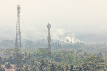 Landscape view of lush palm groves and jungle, with two mobile phone antennas and smoke from some burning in the distance, surrounding the Borobudur temple in Central Java, Indonesia.