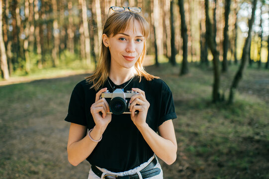 Portrait of beautiful young female photographer with short fair hair in black t-shirt holds a new camera and takes a lot of photos in the big forest in sunny weather