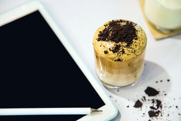 High angle view a cup of Dalgona coffee with cookie crumb next to a white tablet on a white table and cup of milk blurred background.