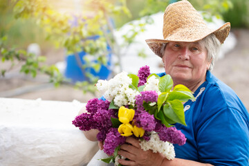 Gray-haired beautiful elderly woman with a bouquet of lilacs outdoors. Pensioner in the country.