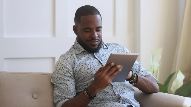 Smiling Black Man Using Digital Tablet Sit On Couch At Home, Young African Guy Holding Computer Looking At Screen Reading E-book App Online Relaxing On Leisure With Wireless Device Work Study Online