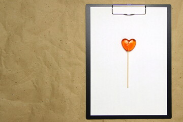 The tablet with a clip for the clip of paper with a white sheet a4 lies against the background of craft brown crumpled paper. On top lies a caramel lollipop of orange red in the shape of a heart. 