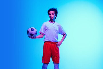 Leader. Football or soccer player on gradient blue studio background in neon light - posing confident with ball. Concept of sport, competition, winning, action, motion, overcoming. Copyspace.