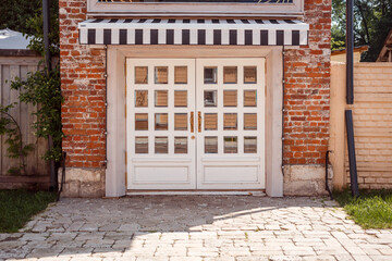 Beautiful exterior, entrance, doors of the shop. Vintage style, white doors with mirror insert and gold-plated door handles. Black and white striped canopy, shade shelter.