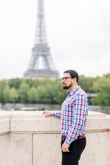 Handsome bearded young man in glasses and casual checkered shirt enjoying wonderful view of Paris and Seine river, standing on the bridge and looking away. Eiffel tower on the background