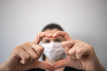 A brunette young woman  in medical face mask  looking at camera and gesturing love sign.