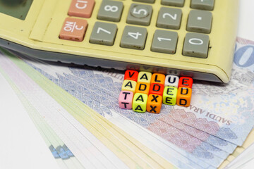 Conceptual value-added tax rollout in Saudi Arabia. Value added tax texts on colorful alphabet beads and calculator over riyals notes. Focus on beads; others in gradient blur.