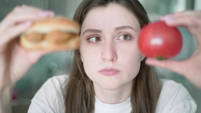Temptation of junk food. The girl is thinking about what to eat. A beautiful woman holds a tomato and cheeseburger and looks at them