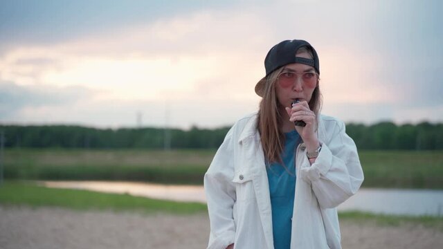 Pretty young girl in cap vape popular ecig gadget vaping deviceat the sunset. Happy brunette vaper girl with e-cig. Portrait of smoker female model with electronic cigarette vaporizer. Ejuice vaping
