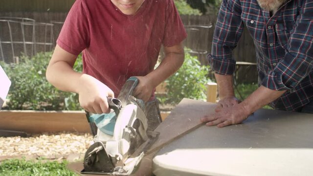 A proud father holds down a piece of wood as his son cuts with a circular saw, they high five