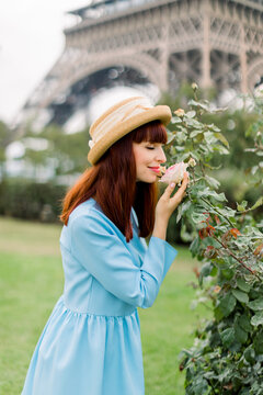 Romantic photo of pretty young french woman with eyes closed enjoying smelling the rose flower, while walking outdoors in Paris, standing in front of Eiffel tower. Red haired girl in hat near roses