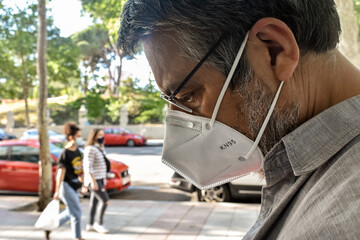 Foreground of a middle-aged man wearing a KN95 (or FFP2) face mask who walks down a city street with the bokeh background of a park and people wearing face masks. “New normality” concept
