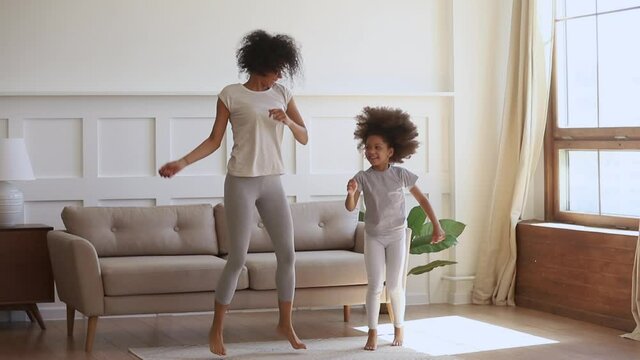 Active happy african mom and cute little kid daughter jumping dancing at home, carefree funny black family mother having fun with small child girl laughing do morning exercise together in living room