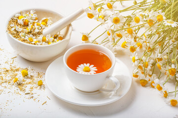 White cup of hot chamomile tea, a mortar with dry daisies buds and a bouquet of Matricaria chamomilla on concrete background. Herbal drink, healthy lifestyle concept.