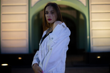 A teenage girl poses against the background of a white and green building arch. Portrait of a brunette at night.