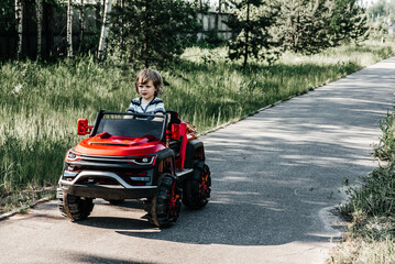 curly-haired boy in a striped T-shirt rides a red big toy car driving on an asphalt path. day off, outdoor recreation