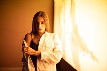 The brunette poses at night in front of a glowing yellow banner. A teenage girl in a white denim jacket, black t-shirt at night illuminated by yellow light.