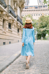 Back view of young stylish red-haired woman in blue dress and hat crossing the street in Paris, walking to Eiffel tower