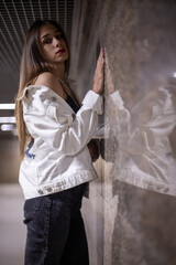 A teenage girl stands facing the wall holding her hand in her reflection. Brunette in the underpass at night, reflection from the wall. A girl in a white denim jacket and black pants. Long hair.
