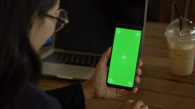 An over the shoulder shot of a young woman video chatting outside with green screen on her smart phone.