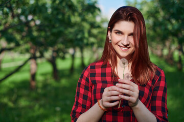 a young girl in a black and red checked shirt with red hair stands on a natural green background in a Park and holds a white dandelion in her hands, looks at it and smiles