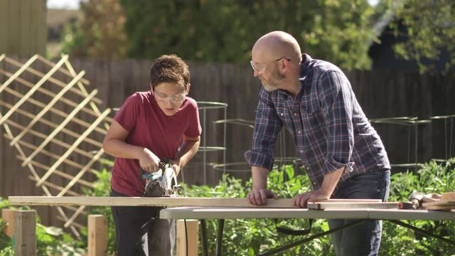 A proud father watches as his son uses a circular saw while working on a wood carpentry project