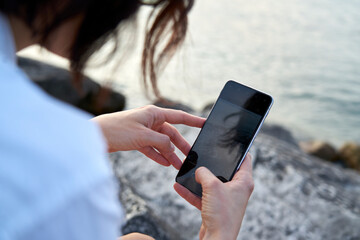 Close-up photograph of a blue screen smartphone held by a woman. In the background the sunset light and the sun reflecting on the blue water of Lake Garda.