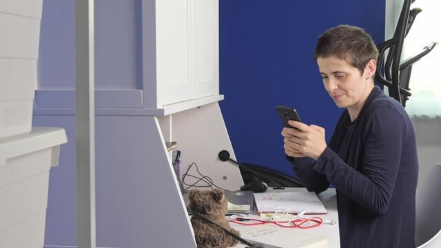 Young woman answers the smartphone while she is in smart working at home in the room, sitting at the desk in front of the laptop